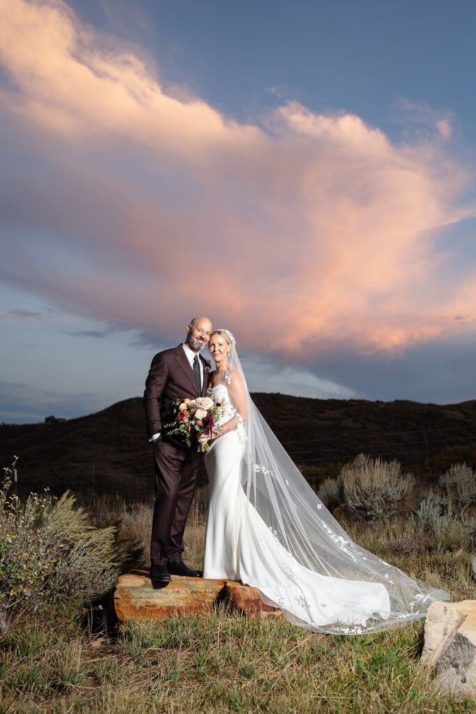 Bride and groom posing beneath dramatic pink sunset clouds with a flowing cathedral veil at High West Distillery in Park City, Utah, during a luxury destination wedding planned by Shellie Ferrer Events.