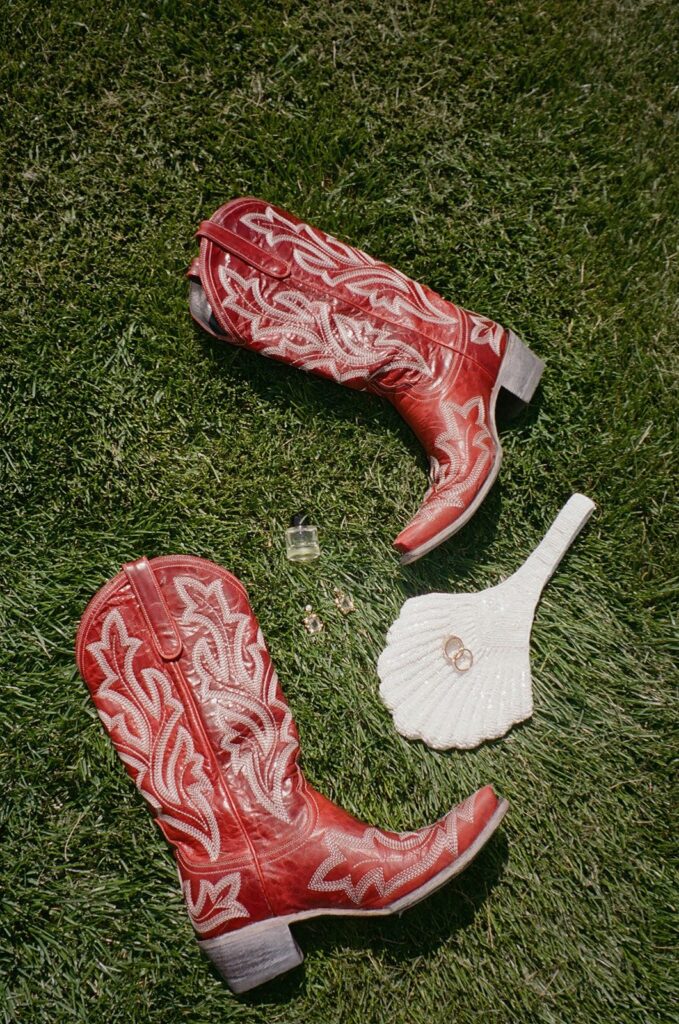 Red embroidered western wedding boots, wedding rings, bridal earrings, perfume, and a white shell-shaped bridal handbag styled on the lawn at a private residence wedding off Wolfe Creek Road in Utah, planned by Shellie Ferrer Events.