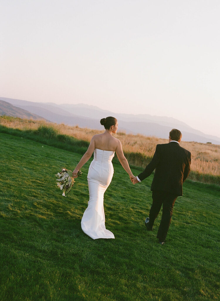 Bride and groom holding hands while walking across a lawn at sunset with mountain views during a destination wedding on Wolfe Creek Ranch Road in Utah, planned by Shellie Ferrer Events.