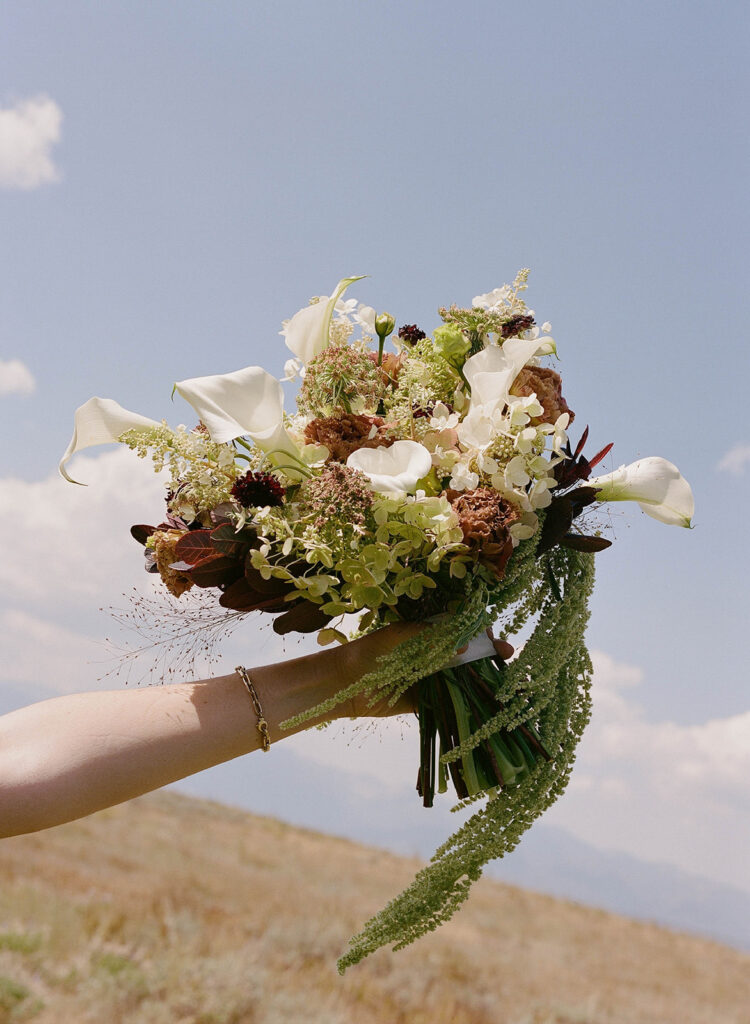 Organic bridal bouquet featuring white calla lilies, hydrangeas, amaranthus, and seasonal blooms held against the Utah sky at a private residence wedding on Wolfe Creek Ranch Road, planned by Shellie Ferrer Events.