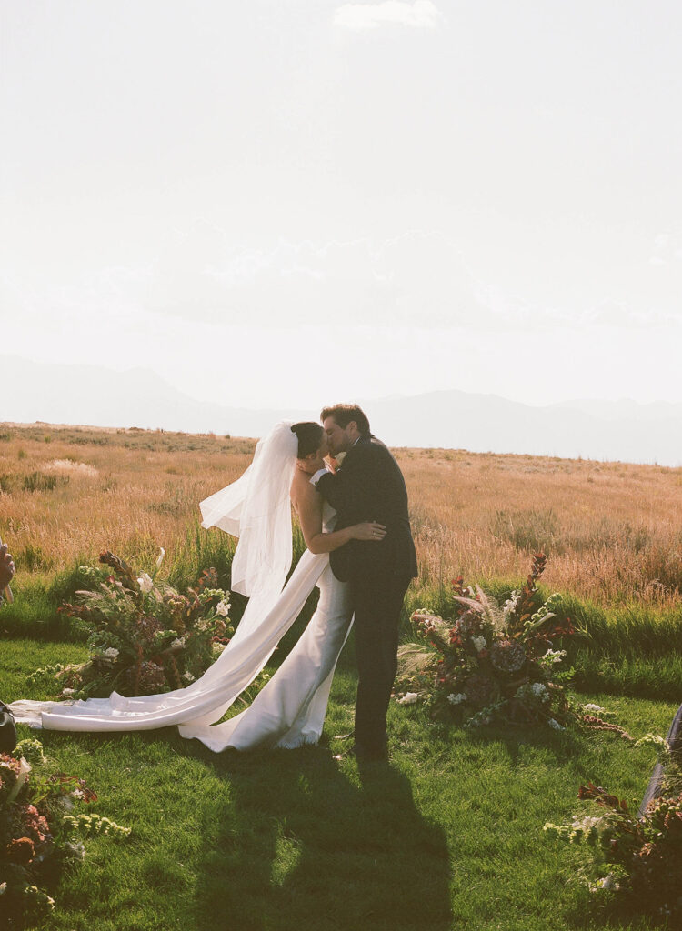 Bride and groom sharing their first kiss at an outdoor destination luxury wedding ceremony surrounded by lush floral arrangements and mountain views on Wolfe Creek Ranch Road in Utah, planned by Shellie Ferrer Events.