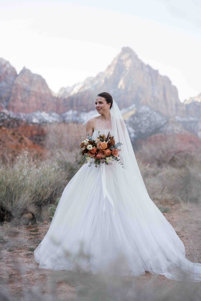 Bride holding a bouquet of flowers standing in Zion National Park, Utah, with red rock cliffs in the background.