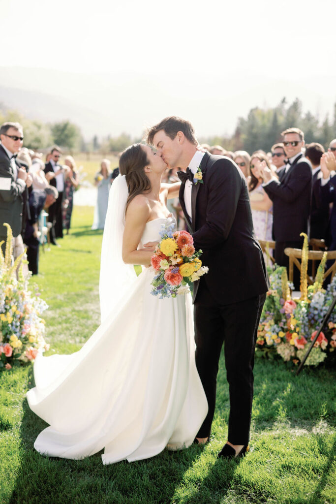 Bride and groom sharing their first kiss during an outdoor wedding ceremony surrounded by guests and lush florals at 4U Ranch in Peoa, Utah, planned by Shellie Ferrer Events.