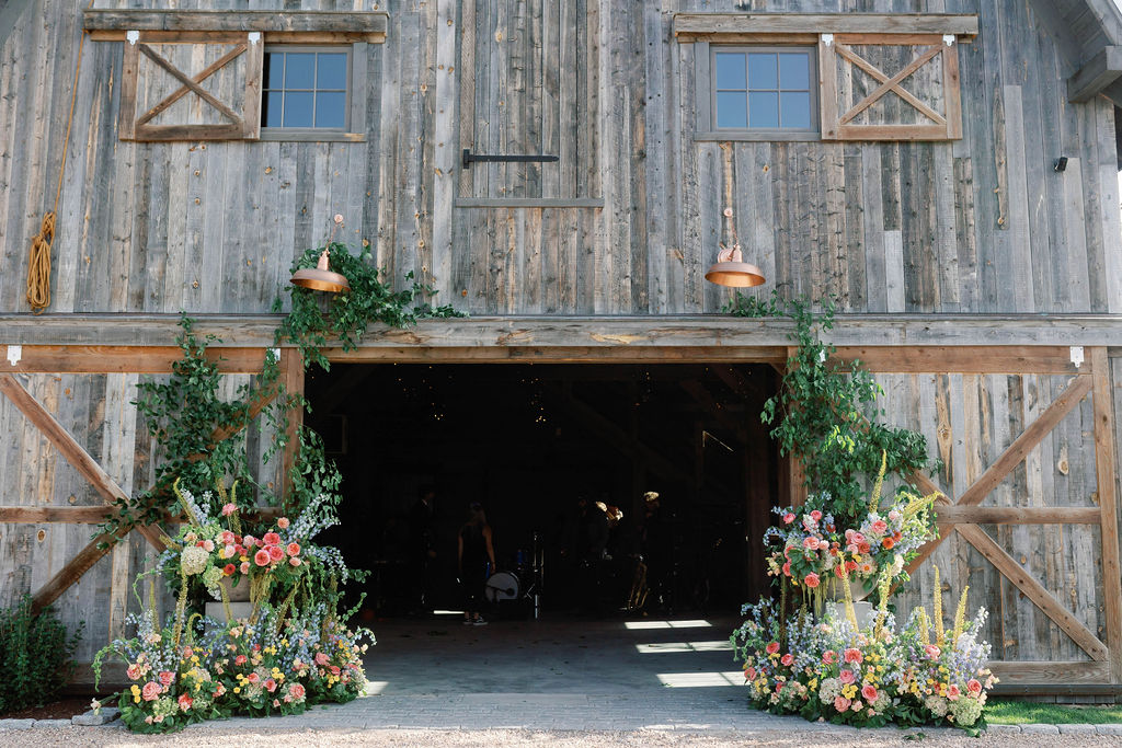 Lush floral installation with colorful garden roses, delphinium, and cascading greenery framing the entrance of a rustic barn at 4U Ranch in Peoa, Utah, planned by Shellie Ferrer Events.