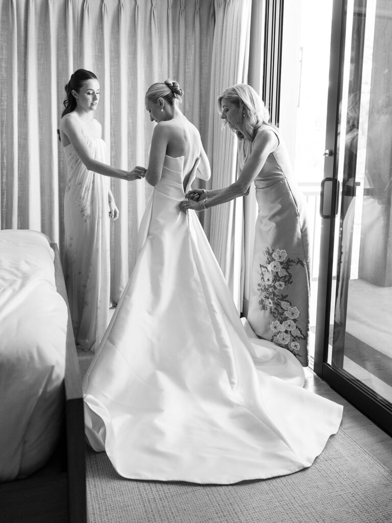 Black-and-white photograph of two women helping a bride fasten her wedding gown at Snowpine Lodge in Little Cottonwood Canyon, Utah.