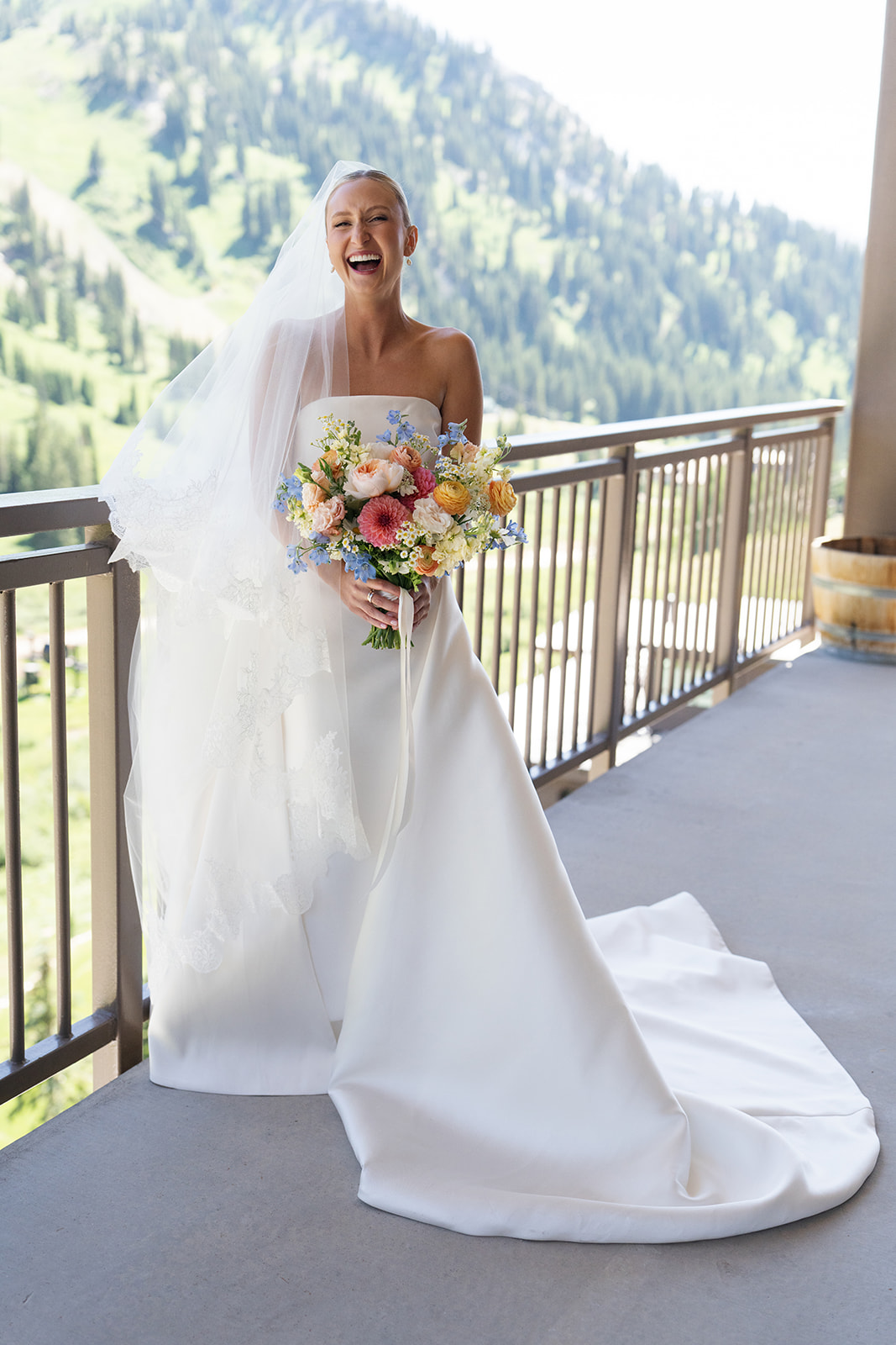 Smiling bride holding a colorful floral bouquet on a balcony overlooking the mountains at Snowpine Lodge in Little Cottonwood Canyon, Utah.